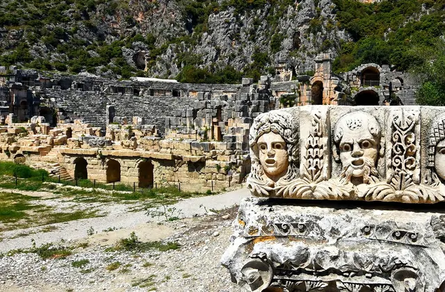 Tres caras de máscaras teatrales talladas adornan un fragmento arquitectónico de piedra caído en primer plano en el sitio arqueológico de Myra, provincia de Antalya, con los muros arqueados en ruinas del teatro romano y el acantilado rocoso extendiéndose al fondo.