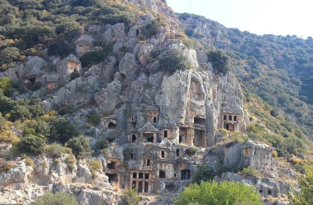 Una vista gran angular del acantilado completo de la necrópolis de Myra, provincia de Antalya, revela docenas de entradas de tumbas licianas talladas en roca distribuidas a diferentes alturas en la ladera de piedra caliza gris pálido, con densa vegetación mediterránea verde cubriendo la pendiente in