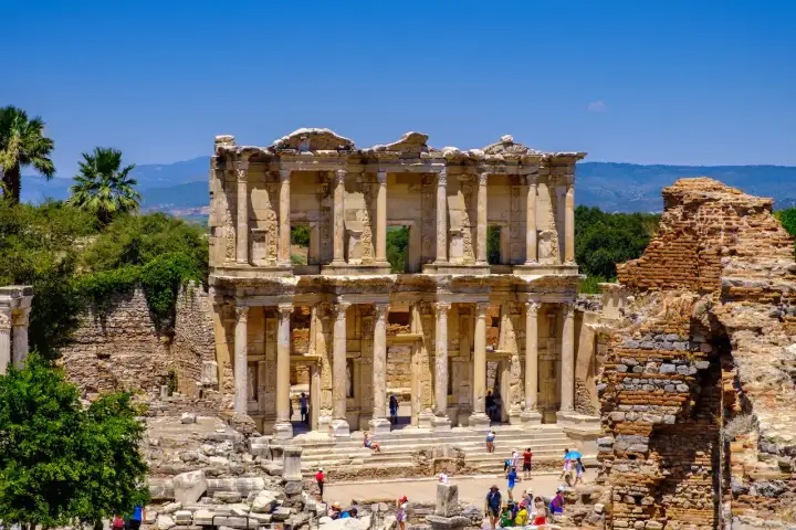 The restored two-storey facade of the Library of Celsus at Ephesus rises against a vivid blue sky, its Corinthian columns and four virtue-statue niches framing tourists who gather on the forecourt steps near Selçuk, Turkey.