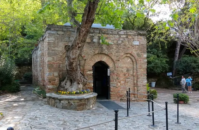The House of the Virgin Mary near Ephesus — a small Byzantine stone chapel with a brick-arched doorway and an ancient gnarled tree growing beside its wall — sits in a wooded hillside clearing above Selçuk, Turkey.