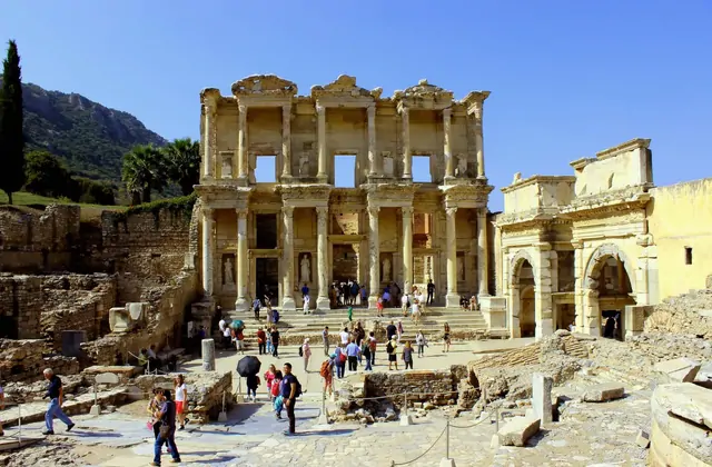 Crowds of visitors explore the sun-drenched forecourt of the Library of Celsus at Ephesus, its two-storey marble facade and the arched Gate of Mazaeus and Mithridates flanking it on the right, with cypress trees and the Panayır Dağ hillside beyond, near Selçuk, Turkey.