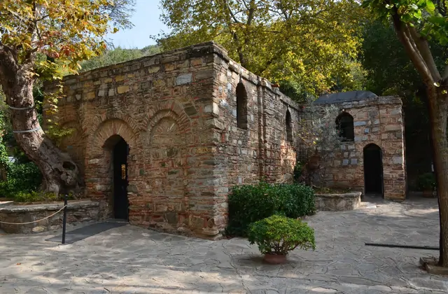 Il tranquillo cortile della Casa della Vergine Maria vicino a Efeso incornicia due strutture in pietra bizantina — la cappella principale e il suo piccolo annesso — circondate da alberi che si tingono d'oro autunnale sui pendii boscosi del Bülbül Dağı sopra Selçuk, in Turchia.