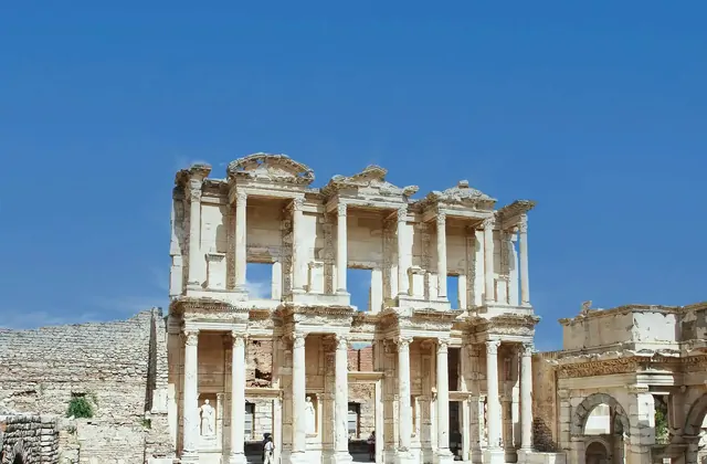 The stark white marble facade of the Library of Celsus at Ephesus stands sharply against a cloudless deep-blue sky at midday, its two tiers of Corinthian columns and decorative pediments clearly visible with barely a visitor in sight near Selçuk, Turkey.