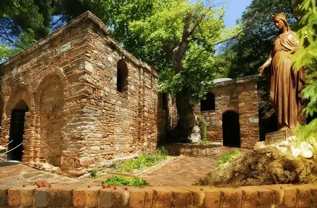 A bronze statue of the Virgin Mary stands in the lush garden beside the ancient stone chapel of the House of the Virgin Mary near Ephesus, Turkey, where summer foliage wraps the Byzantine walls in vibrant green on the wooded slopes of Bülbül Dağı.