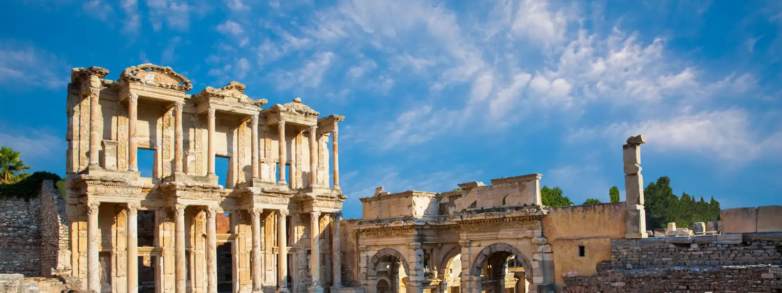 The Library of Celsus and the Gate of Mazaeus and Mithridates stand side by side in the morning light at Ephesus, Turkey, their Roman masonry bathed in warm gold under a theatrical blue sky with wispy clouds, the marble forecourt stretching empty before them.