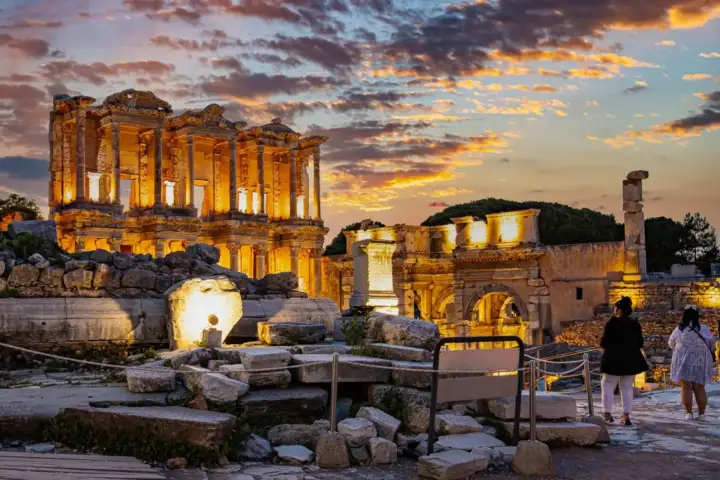 The Library of Celsus at Ephesus blazes with warm amber floodlight at dusk, its ornate columns and the Gate of Mazaeus glowing against a fiery orange-pink sunset sky above the ancient city near Selçuk, Turkey, as two visitors watch from the foreground.