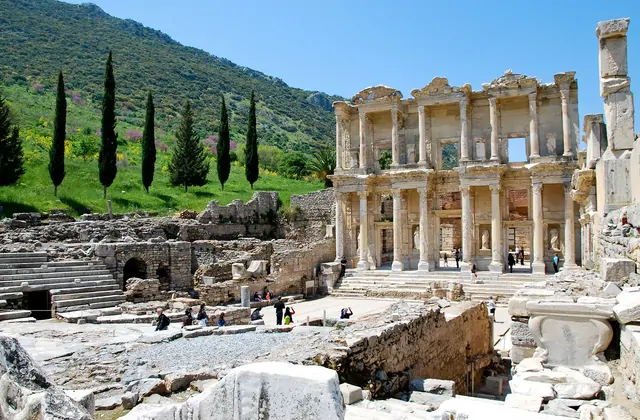 The Library of Celsus rises above a wide marble forecourt at Ephesus, framed by tall green cypress trees on the Panayır Dağ hillside to the left, the Gate of Mazaeus visible to the right under a bright blue sky near Selçuk, Turkey.