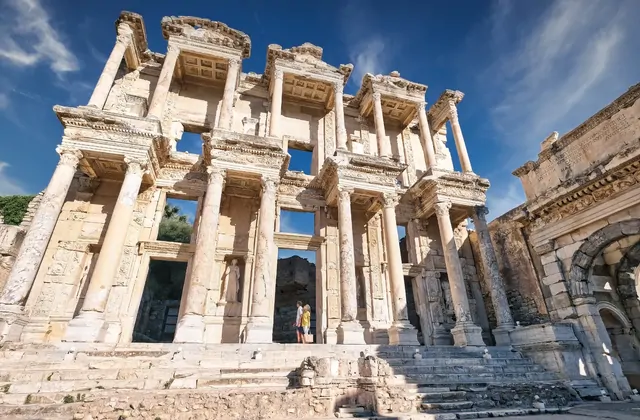 A low, wide-angle view looking up at the Library of Celsus at Ephesus emphasises the monumental scale of its two-storey Corinthian facade, with a single visitor in a yellow jacket dwarfed at its base and a sky of blue and white clouds above, Turkey.