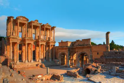 Warm sunrise light bathes the entire two-storey facade of the Library of Celsus at Ephesus in deep amber, turning its marble columns and carved pediments a rich burnt-orange while the Gate of Mazaeus glows behind to the right, near Selçuk, Turkey.