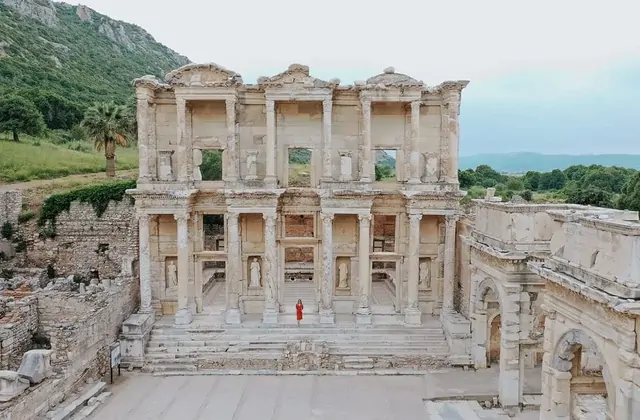 A lone visitor in a red dress stands centred on the grand staircase of the Library of Celsus at Ephesus in this elevated view, the full two-storey facade with its four virtue-statue niches perfectly symmetrical, with the Gate of Mazaeus to the right and a green hillside behind, Turkey.