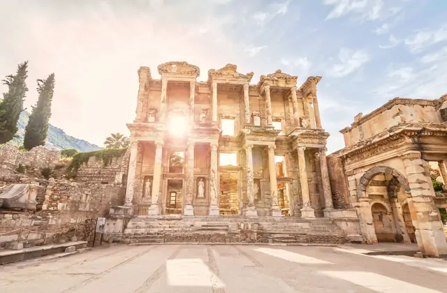Sunlight bursts dramatically through the central window opening of the Library of Celsus at Ephesus, creating a starburst effect against the two-storey marble facade, with cypress trees flanking the ruins and a hazy sky above, near Selçuk, Turkey.