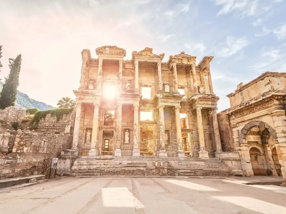 Sunlight bursts dramatically through the central window opening of the Library of Celsus at Ephesus, creating a starburst effect against the two-storey marble facade, with cypress trees flanking the ruins and a hazy sky above, near Selçuk, Turkey.