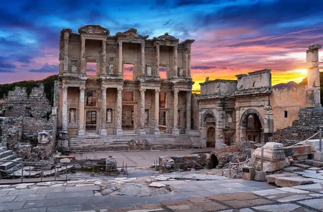 Deep twilight descends on the Library of Celsus and the Gate of Mazaeus at Ephesus, the warm floodlit stone facades contrasting with a deep-blue and orange horizon sky while the ancient forecourt falls into evening shadow near Selçuk, Turkey.