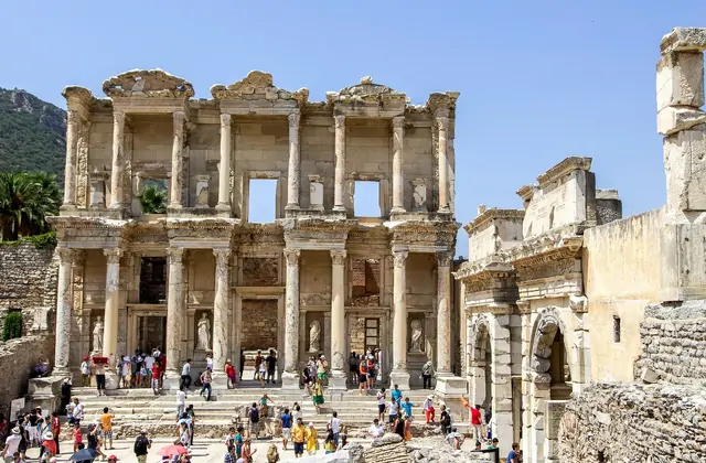 A wide midday view of the Library of Celsus and the Gate of Mazaeus at Ephesus shows scattered visitors crossing the open forecourt under a blue sky dotted with white clouds, the forested Panayır Dağ hillside rising behind the ancient monument near Selçuk, Turkey.