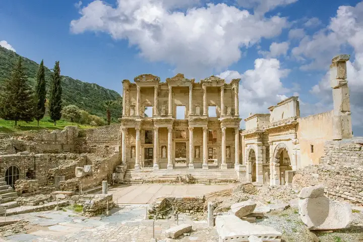 The Library of Celsus stands centred in a wide landscape composition at Ephesus, the Gate of Mazaeus and Mithridates to its right, tall cypress trees and a forested hillside behind, and a partly cloudy sky above, photographed near Selçuk, Turkey.