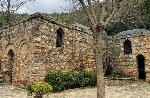The House of the Virgin Mary near Ephesus in winter, its ancient stone chapel framed by a bare-branched tree in the foreground and a votive candle stand visible to the right, the tranquil pilgrimage site enclosed by the wooded hillside of Bülbül Dağı, Turkey.
