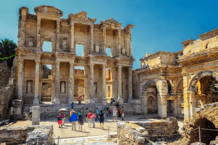 Tourists mill around the base of the Library of Celsus at Ephesus in this close medium-shot, with the Gate of Mazaeus and Mithridates to the right under a cloudless summer-blue sky, the monument's Corinthian columns and arched niches sharp in the clear light near Selçuk, Turkey.