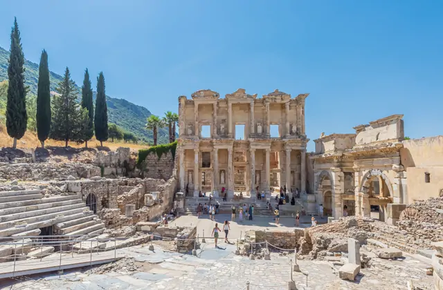 Un'ampia ripresa angolata della Biblioteca di Celso a Efeso, scattata accanto alle rovine del teatro antico in basso a sinistra, mostra le colline ricoperte di cipressi dietro il monumento e la Porta di Mazeo a destra in una chiara giornata estiva vicino a Selçuk, in Turchia.