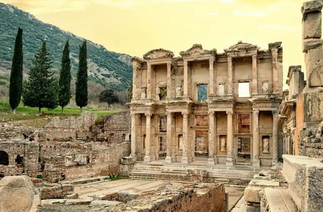Altos cipreses en la ladera del Panayır Dağ enmarcan la Biblioteca de Celso en Éfeso bañada en cálida luz de hora dorada, la Puerta de Mazeo brillando a la derecha y ruinas arqueológicas dispersas rodeando el monumento cerca de Selçuk, Turquía.