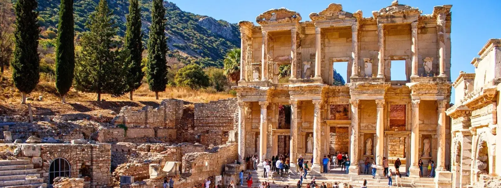 Visitors crowd the steps and forecourt of the Library of Celsus at Ephesus on a sunny autumn day, the two-storey marble facade rising above the bustle with tall cypress trees and the Panayır Dağ mountain range stretching across the background near Selçuk, Turkey.