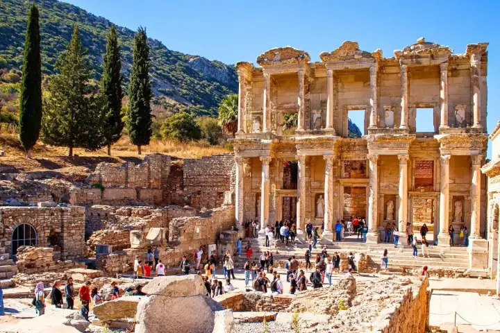 Visitors crowd the steps and forecourt of the Library of Celsus at Ephesus on a sunny autumn day, the two-storey marble facade rising above the bustle with tall cypress trees and the Panayır Dağ mountain range stretching across the background near Selçuk, Turkey.