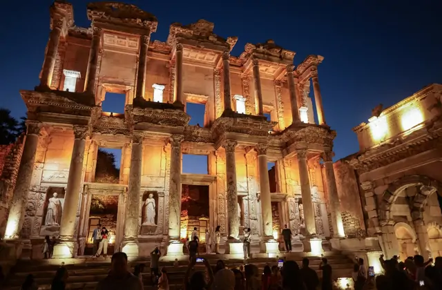 The Library of Celsus at Ephesus glows with dramatic amber floodlighting against a deep night sky, crowds of visitors photographing on the lit steps and forecourt while the Gate of Mazaeus blazes to the right in this spectacular nocturnal scene near Selçuk, Turkey.