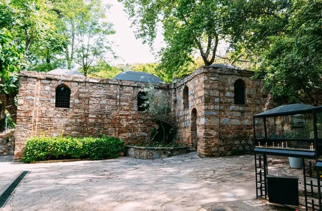 The House of the Virgin Mary near Ephesus in full summer foliage, the stone chapel and its smaller annex visible side by side in the courtyard with a wish-tying stand and votive candle holder to the right, enclosed by the dense trees of Bülbül Dağı hillside, Turkey.