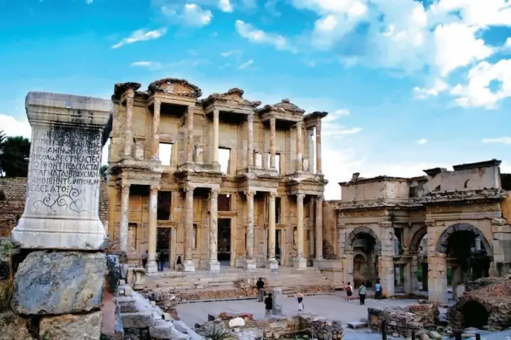 An ancient inscribed stele in the foreground frames the Library of Celsus at Ephesus, its Greek lettering still legible, while the Gate of Mazaeus stands to the right and warm afternoon light gilts the limestone masonry of this UNESCO World Heritage site near Selçuk, Turkey.