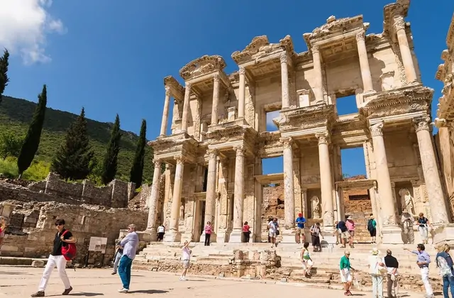 Turisti vestiti di colori vivaci riempiono il luminoso cortile in marmo della Biblioteca di Celso a Efeso in una trafficata giornata estiva, la facciata a due piani che si innalza sulle folle contro un cielo azzurro limpido, con i cipressi che risalgono il versante dietro, vicino a Selçuk, in Turchi