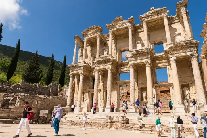 Colourfully dressed tourists fill the sun-bright marble forecourt of the Library of Celsus at Ephesus on a busy summer day, the two-storey facade towering above the crowds against a clear blue sky, with cypress trees climbing the hillside behind, near Selçuk, Turkey.