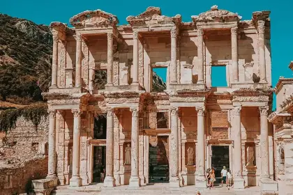 The Library of Celsus at Ephesus photographed in a compact frontal shot under a vivid turquoise-blue sky, with the rocky Panayır Dağ summit visible just above the roofline and only a handful of visitors at the base in the bright sunshine near Selçuk, Turkey.