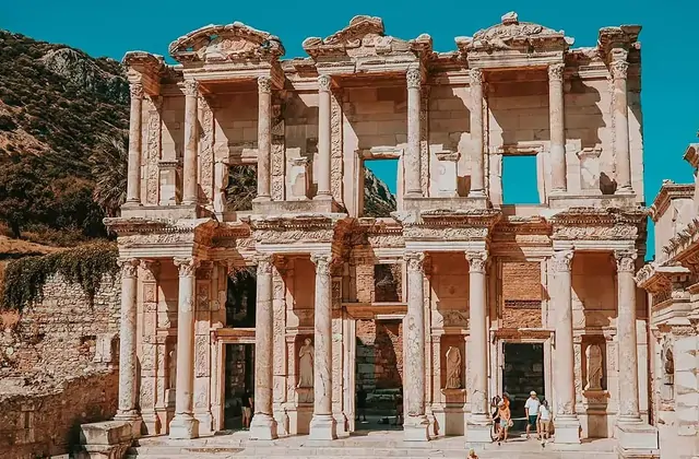 The Library of Celsus at Ephesus photographed in a compact frontal shot under a vivid turquoise-blue sky, with the rocky Panayır Dağ summit visible just above the roofline and only a handful of visitors at the base in the bright sunshine near Selçuk, Turkey.