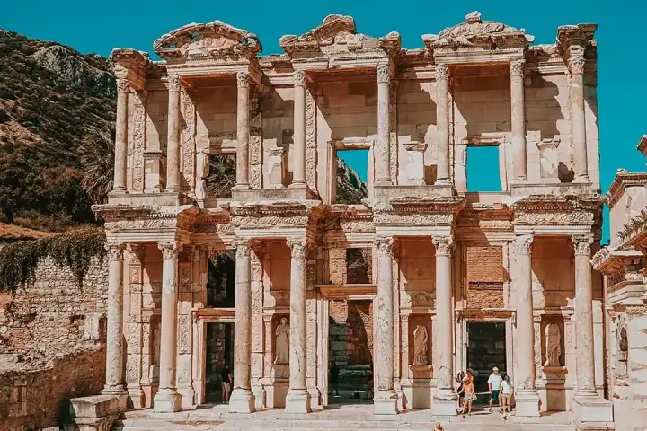 The Library of Celsus at Ephesus photographed in a compact frontal shot under a vivid turquoise-blue sky, with the rocky Panayır Dağ summit visible just above the roofline and only a handful of visitors at the base in the bright sunshine near Selçuk, Turkey.