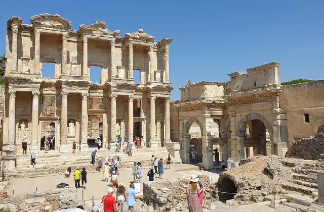 Visitors stream across the open forecourt of the Library of Celsus at Ephesus on a busy midday visit, the Gate of Mazaeus and Mithridates visible to the right, the full two-storey facade and the adjacent Roman arch forming an iconic ensemble near Selçuk, Turkey.