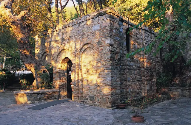 Late-afternoon golden light warms the stone facade of the House of the Virgin Mary near Ephesus, long tree shadows falling across the paving stones of the quiet courtyard on the wooded slopes of Bülbül Dağı above Selçuk, Turkey.