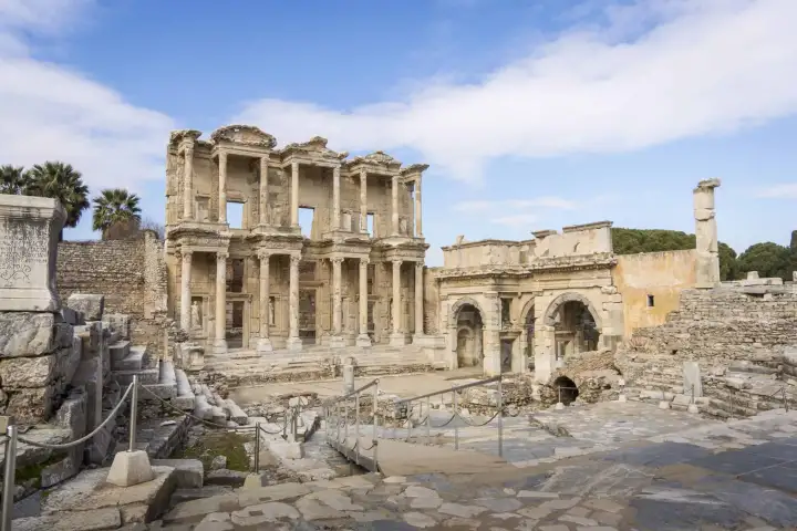 The Library of Celsus and the Gate of Mazaeus at Ephesus stand in clear winter light under a blue sky with scattered clouds, the bare hillside behind the monument and virtually no visitors in the open forecourt near Selçuk, Turkey.