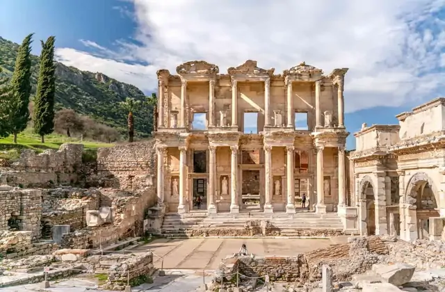 A sweeping panoramic view of the Library of Celsus at Ephesus in spring, the Gate of Mazaeus to the right and the marble forecourt stretching in the foreground, the green Panayır Dağ hillside and a palm tree providing a lush backdrop under a bright overcast sky, Turkey.