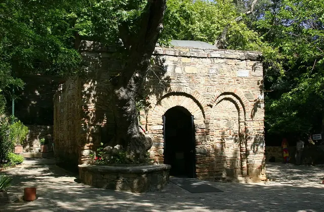 La Casa della Vergine Maria vicino a Efeso è semi-nascosta in estate dalla chioma protesa di un antico albero, con il portale in pietra ad arco intravisto attraverso l'ombra screziata sul sacro versante del Bülbül Dağı vicino a Selçuk, in Turchia.