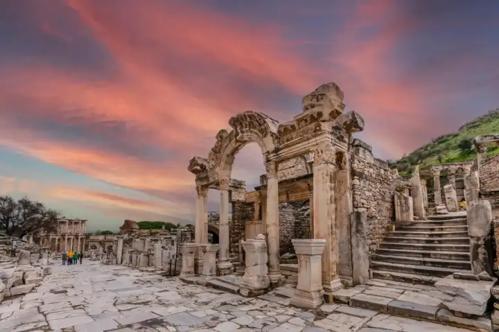 The Temple of Hadrian on Curetes Street at Ephesus glows under a spectacular pink-and-purple sunset sky, the ornate carved arch in the foreground with the marble-paved Sacred Way stretching towards the distant Library of Celsus visible in the background, near Selçuk, Turkey.