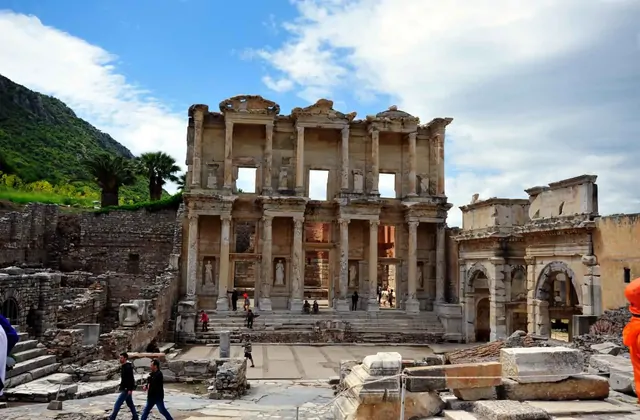 La Biblioteca de Celso en Éfeso vista en un nublado día de primavera frente a la brillantemente verde ladera boscosa del Panayır Dağ, con visitantes cruzando el atrio de mármol y la Puerta de Mazeo a la derecha en esta vista paisajística gran angular cerca de Selçuk, Turquía.