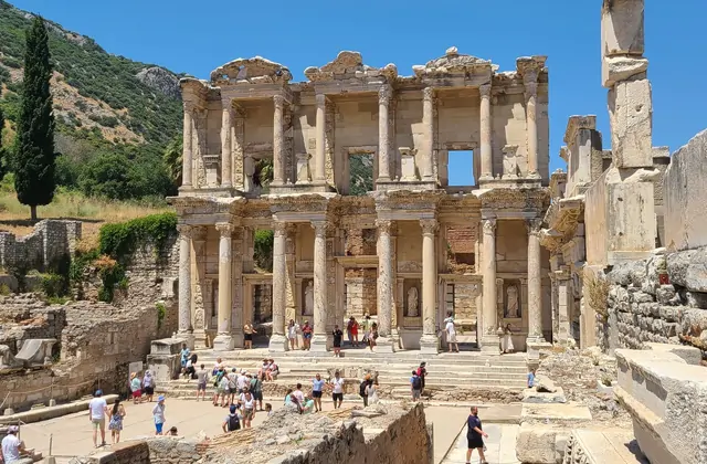 Visitors crowd the steps of the Library of Celsus at Ephesus on a clear summer day, the two-storey marble facade with its Corinthian columns and carved pediments rising sharply against a bright blue sky, the Panayır Dağ mountain visible behind near Selçuk, Turkey.