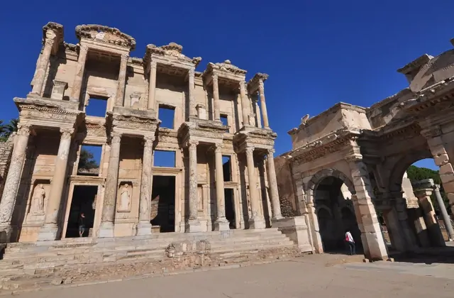 Una vista amplia y baja de la Biblioteca de Celso y la Puerta de Mazeo en Éfeso bajo un cielo azul profundo y despejado, los dos monumentos antiguos enmarcando el vacío atrio de mármol con solo un par de visitantes visibles, resaltando la escala monumental de la Éfeso romana, Turquía.