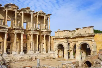 The Library of Celsus and the Gate of Mazaeus at Ephesus form a dramatic wide-angle panorama in pale winter light, the adjacent Roman walls and arcade visible to the left, while a warm yellowish sky crowns the ancient monument near Selçuk, Turkey.