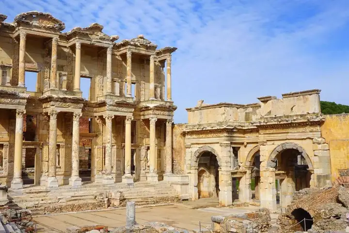The Library of Celsus and the Gate of Mazaeus at Ephesus form a dramatic wide-angle panorama in pale winter light, the adjacent Roman walls and arcade visible to the left, while a warm yellowish sky crowns the ancient monument near Selçuk, Turkey.