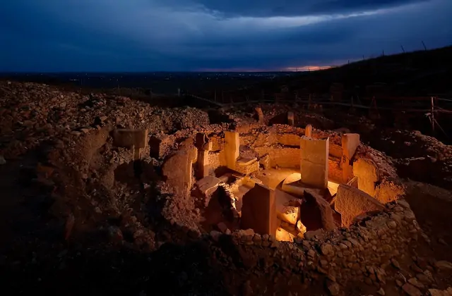 Göbekli Tepe's excavated circular enclosure glows under warm amber floodlights at dusk, with T-shaped limestone pillars casting long shadows inside the stone-walled ritual chamber while the distant city lights of Şanlıurfa shimmer on the dark horizon.