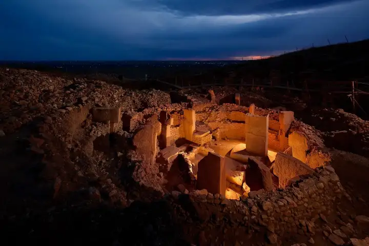 Göbekli Tepe's excavated circular enclosure glows under warm amber floodlights at dusk, with T-shaped limestone pillars casting long shadows inside the stone-walled ritual chamber while the distant city lights of Şanlıurfa shimmer on the dark horizon.