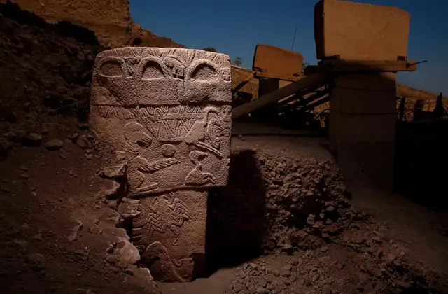 A close-up night view of a Göbekli Tepe limestone pillar at the excavation site reveals dense, stacked relief carvings including birds, serpents, and geometric patterns covering the entire front face, with a second T-shaped pillar looming in the dark background.