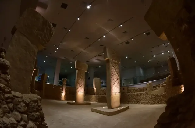 Inside the Şanlıurfa Museum, a full reconstructed Göbekli Tepe enclosure fills a dimly lit gallery hall, with multiple tall T-shaped pillars arranged in a circle around a central pair, all positioned within rebuilt dry-stone circular walls under recessed ceiling spotlights.