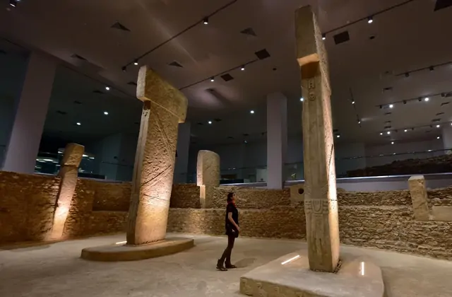 A visitor gazes upward at a towering T-shaped Göbekli Tepe pillar replica inside the Şanlıurfa Museum, the pillar's shaft bearing a carved snake motif lit from below, while additional T-shaped pillars of varying heights stand in a circle within reconstructed dry-stone walls under bright track lighti
