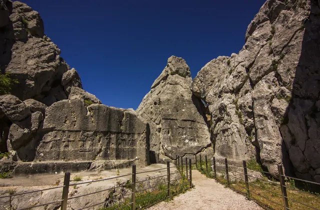 La Camera B di Yazılıkaya vicino a Hattusa, Boğazkale, Turchia, forma un angusto corridoio roccioso dove un fregio scolpito di divinità ittite in cammino riempie la parete rocciosa destra, affiancato da ringhiere metalliche che guidano i visitatori moderni lungo il sacro passaggio.